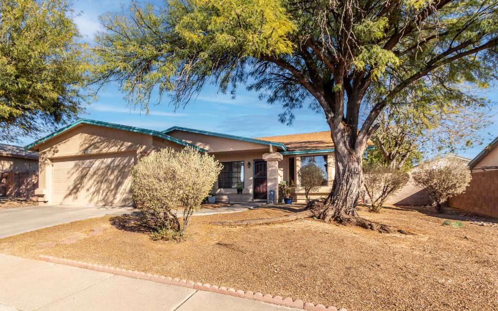 Tucson east side home for sale, beige frame stucco house with a 2 car garage and large mesquite tree and desert shrubs in front yard.