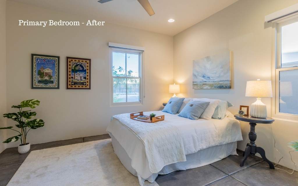 A primary bedroom staged with an area rug, a bed, nightstands, art on the walls, table lamps, and a plant. 