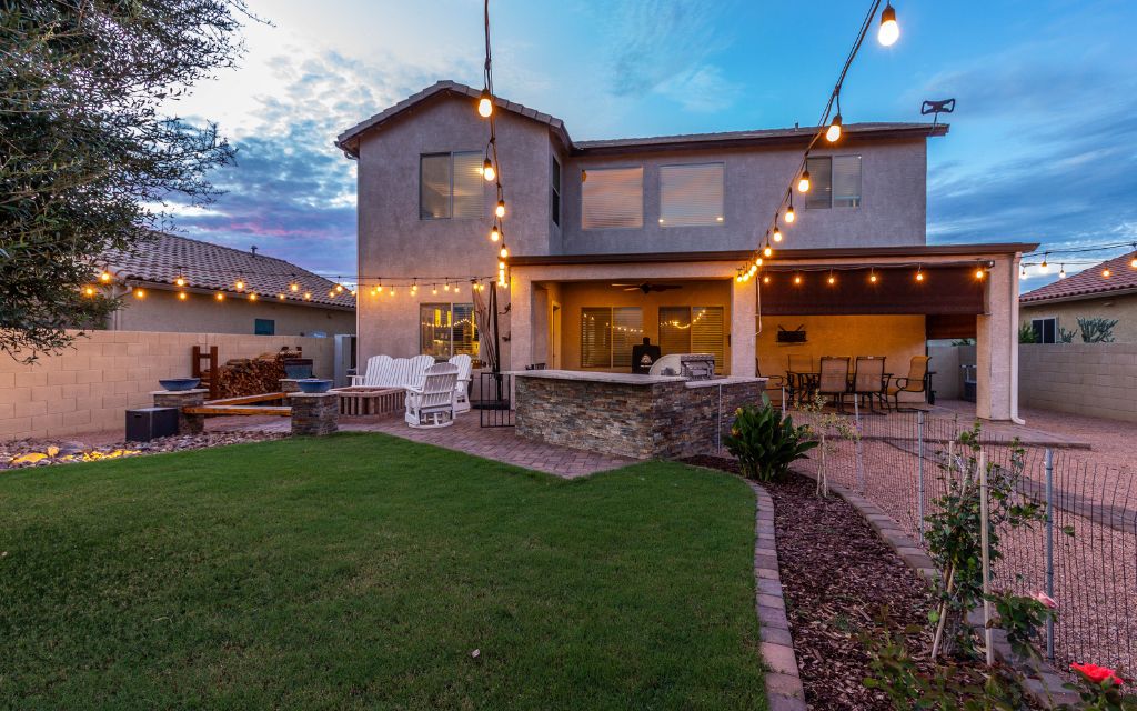 backyard of a 2-story home in Red Rock, Arizona with a lawn, string lights, a built-in grill, covered patio with a table and chairs and a uncovered patio with chairs around a firepit.