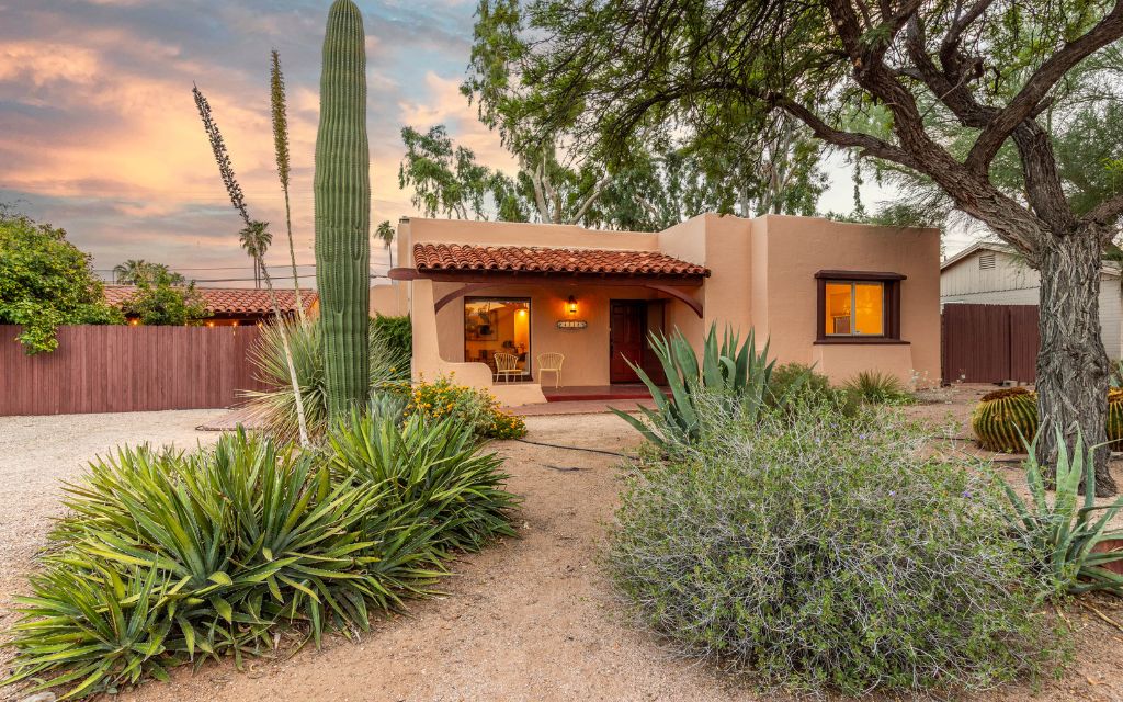Spanish Colonial Revival style home with peach painted stucco and inviting front porch with a tile roof, featuring mature desert landscaping in the front yard.